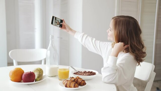 A side view of the smiling little girl taking a selfie photo on the phone and showing different gestures at the breakfast