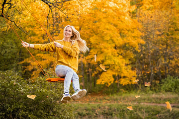 Happy young blonde woman with loose hair smiles and rides on a rope swing in autumn in the park.