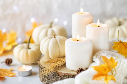 Hello Autumn or Happy Thanksgiving concept. White burning candles with white pumpkins at the background, knitted plaid and autumnal leaves. Shallow depth of field