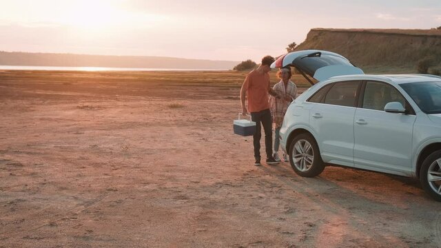 Beautiful Multinational Couple Taking Out A Picnic Basket From The Trunk Of A Car And Going To A Picnic