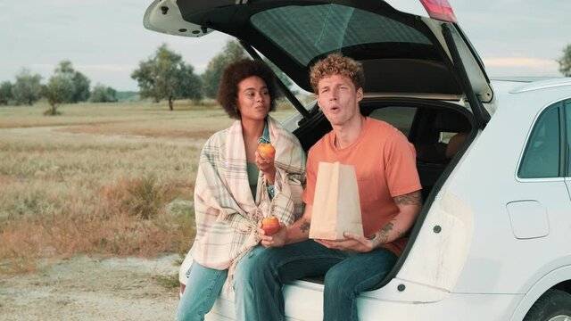 Multinational couple eating peaches while sitting in the trunk of a car in nature