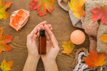 Young adult woman hands holding brown shower lotion bottle. Fresh colorful maple leaves, soap bar and bath ball on wooden table background. Closeup. Care about body skin in autumn time. Top down view.