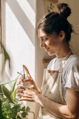 Young happy mixed race woman is spraying houseplants with water from a sprayer at home.Home gardening.Hobby concept.Biophilia design and urban jungle concept.