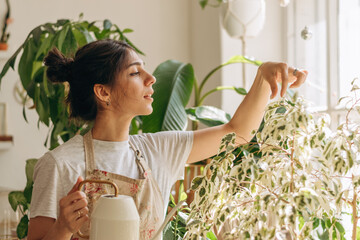 Positive young beautiful mixed race woman in apron is watering houseplants at home.Home gardening.Hobby concept.Biophilia design and urban jungle concept. © Tatyana