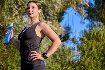 Fitness athlete a young woman listens to music with headphones, drinks water from a reusable soft sports water bottle after training on a sunny day in the park