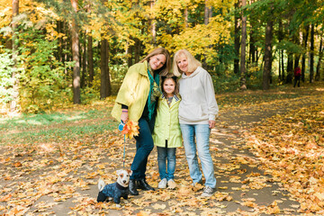 Naklejka na ściany i meble Portrait of three generations of happy beautiful woman and dog looking at camera, hugging and smiling in autumn nature.