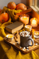 Mug of cocoa or hot chocolate with marshmallows next to the window with candles, pumpkins, book and warm blanket. Cozy home atmosphere in rainy autumn day. Нygge lifestyle concept.