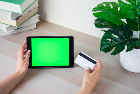 Female hands hold tablet computer with green screen and credit card. Woman sits at table with books. Concept of online shopping and communication technology.