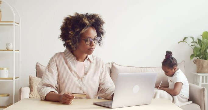 Young african american single mom in glasses, paying online with credit card and laptop, buying something for her daughter on ecommerce website
