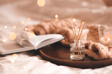 Home perfume in glass bottle with wood sticks, scented burn candles, open paper book and knit wool textile on tray in bedroom close up. Aromatherapy cozy atmosphere lifestyle. Winter warm xmas season
