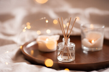 Home perfume in glass bottle with wood sticks, scented burn candles  tray in bedroom close up. Aromatherapy cozy atmosphere lifestyle. Winter warm xmas season. - obrazy, fototapety, plakaty