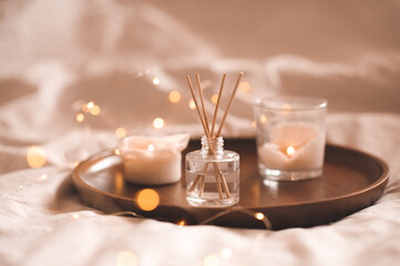 Home perfume in glass bottle with wood sticks, scented burn candles on tray in bedroom close up...