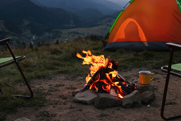 Metal mug with drink and beautiful bonfire near camping tent in mountains