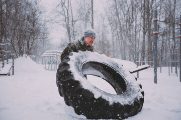 A man lifts a tire during a training session in the park in winter in a snowfall. a man performs the Tire turning exercise in a heavy snowstorm