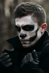 The image of a skeleton on Halloween in the park. Male image for Halloween. A man in the image of Dracula in a black coat and with white makeup. He holds the collar of coat and looks angrily at camera