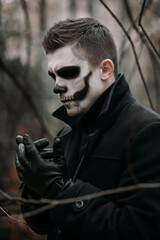 The image of a skeleton on Halloween in the park. Male image for Halloween. A man in the image of Dracula among the bare trees in an autumn park. He is wearing a black coat and leather gloves