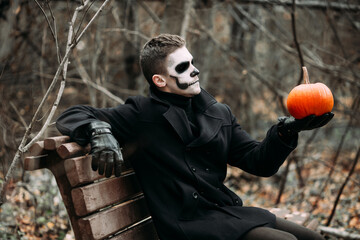 The image of a skeleton on Halloween in the park. Male image for Halloween. A man in a black coat is sitting on a bench and holding a pumpkin at arm's length. There is a park and bare trees