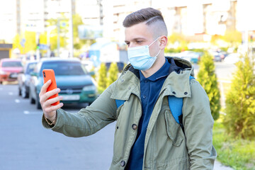 Fototapeta na wymiar Young man in a protective mask makes a video call and talks on a smartphone while standing in the city. Communication between people at a distance during quarantine and pandemic.