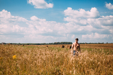 Naked to the torso, a man walks through a field of wheat. The sky is blue with white clouds. The man is wearing denim shorts. The field goes beyond the horizon. Lots of golden spikelets