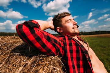 A man in a red plaid shirt leans on a haystack with his hand. Hands behind his head, eyes closed, he smiles. The shirt is open, the naked torso is visible. It's summer outside. A straw in the teeth