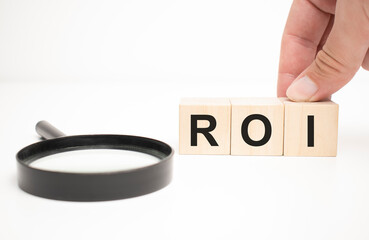 roi text wooden cube blocks and hand holding magnifying glass on table background.