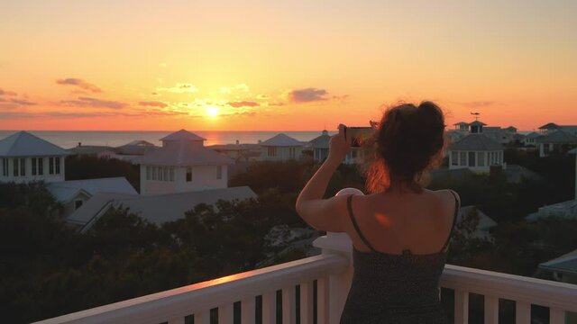 View on colorful sunset with young woman watching photographing with phone Gulf of Mexico in Seaside, Florida on wooden rooftop terrace building house balcony