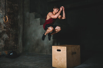The jock jumps onto the cube. The athlete hovered in the air above the cube. Functional crossfit training. High powerful jump. The man is wearing a burgundy T-shirt and black shorts.