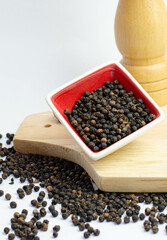 Black pepper in red ceramic bowl on kitchen board, with pepper mill behind, on white background....