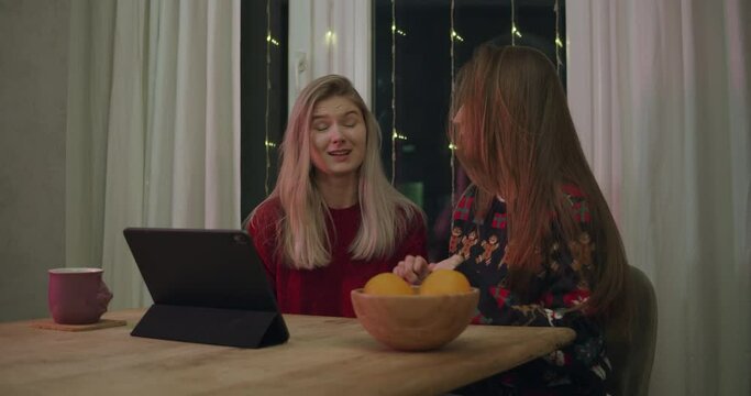 Young women at Xmas using tablet video call. Young girls laugh and talk. Christmas lights decorated with ornament pullovers in dining room at home.