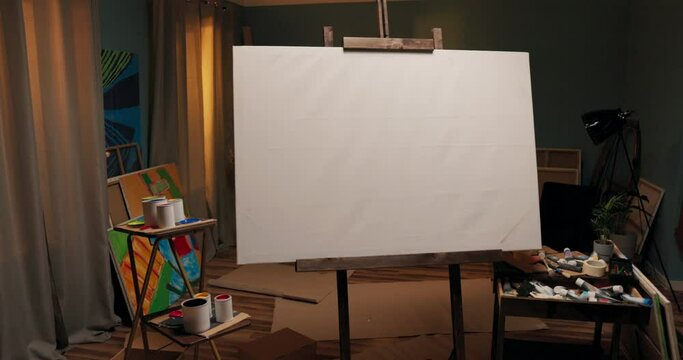 A workspace, a studio of a painter, an artist with a large easel with a canvas in the middle of the room. Around the canvas scattered paints, brushes, cardboard boxes, a mess.