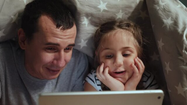 dad and daughter under the blanket with a digital tablet. kid dream online night video games at concept. dad and daughter watching online video under covers with digital tablet. social media