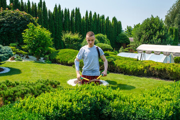 Young male with amputated arm and a prosthesis trimming bushes in a garden in his yard with large...