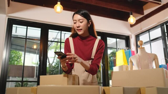 Asian woman use mobile phone to take a picture of banner on top of many boxes during process of delivery product to customer for small business system.