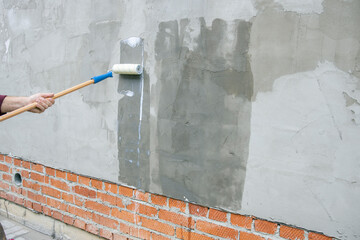 A worker applies the primer to the wall with a long-handled roller. A man's hand rolls a primer...