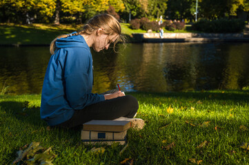 Fototapeta premium A student girl is sitting in the park on the grass and making notes. A stack of books.