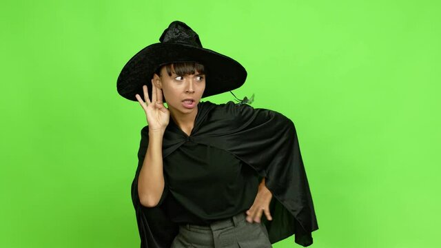 Young woman wearing witch hat for halloween parties listening to something by putting hand on the ear over isolated background. Green screen chroma key
