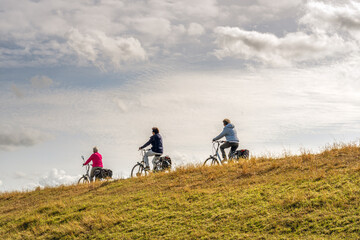 Obraz premium Three unidentified women cycle one behind the other with their electric bicycles on a Dutch dike. It is a sunny day in the autumn season. The grass on the embankment of the dike is yellow and withered
