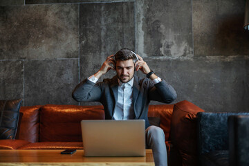 Fototapeta premium Businessman sitting on a couch and putting on headphones. He is preparing for the conference call. Telecommunications, technologies, business meeting, online meeting