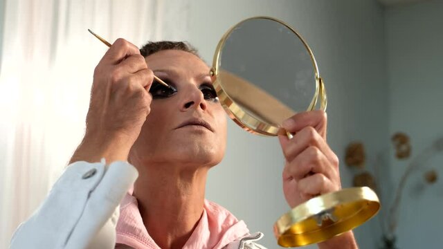 Caucasian male is applying the false eyelashes  looking at the mirror. travesty is doing make up. LGBT