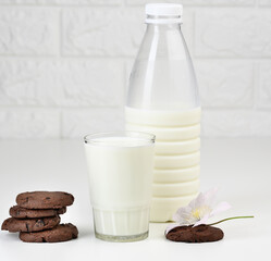 a full glass cup of milk and a clear plastic bottle with milk, next to a stack of round chocolate chip cookies on a white table