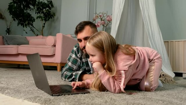 Caucasia family dad and child daughter using laptop computer at home lying on floor, father helps kid girl with homework having fun looking at pc screen doing online shopping, study in internet