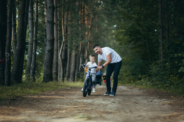 man father teaches little son son riding a bike in the woods on a walk