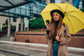 Beautiful woman using a smartphone and holding a yellow umbrella while walking in the city on a rainy day - obrazy, fototapety, plakaty