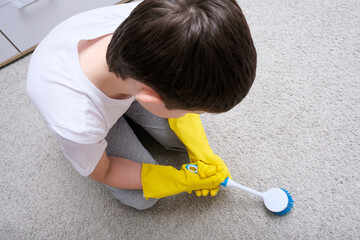A boy in yellow rubber gloves helping his mom and cleaning carpet with a brush, housekeeping and...