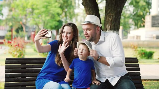 Happy family mom dad son, little baby are photographed using modern smartphone in park in summer. Father toddler mother, parents have fun using smartphone, selfie or video calling together outdoors