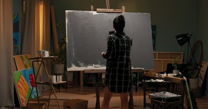 A woman dressed in a checkered shirt walks away from the paint container with a roller soiled in gray, adding a second coat of primer in some places, precise movements