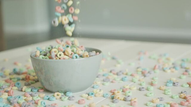 Breakfast cereals in the form of colorful corn rings fall and fill the bowl on the table. Food close-up on the background of scattered flakes. Colored corn flakes and dairy products. Slow motion. High