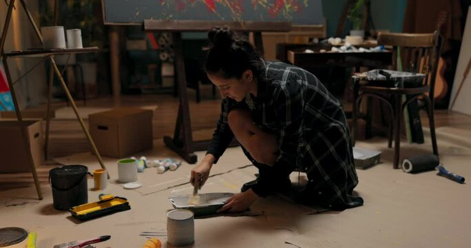 A talented woman is crouch,working in her studio, school, and art class on a new piece of painting related to abstractionism, modernity. The girl crouches on the floor reaching for paint.