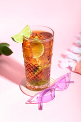 Glass of refreshing Long Island iced tea with lime and stylish sunglasses on color background