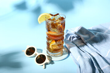 Glass of refreshing Long Island iced tea, sunglasses and towel on color background